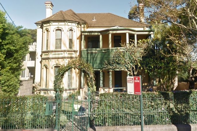 A two-storey house is pictured from the street. There is a green fence and hedging at the perimeter.