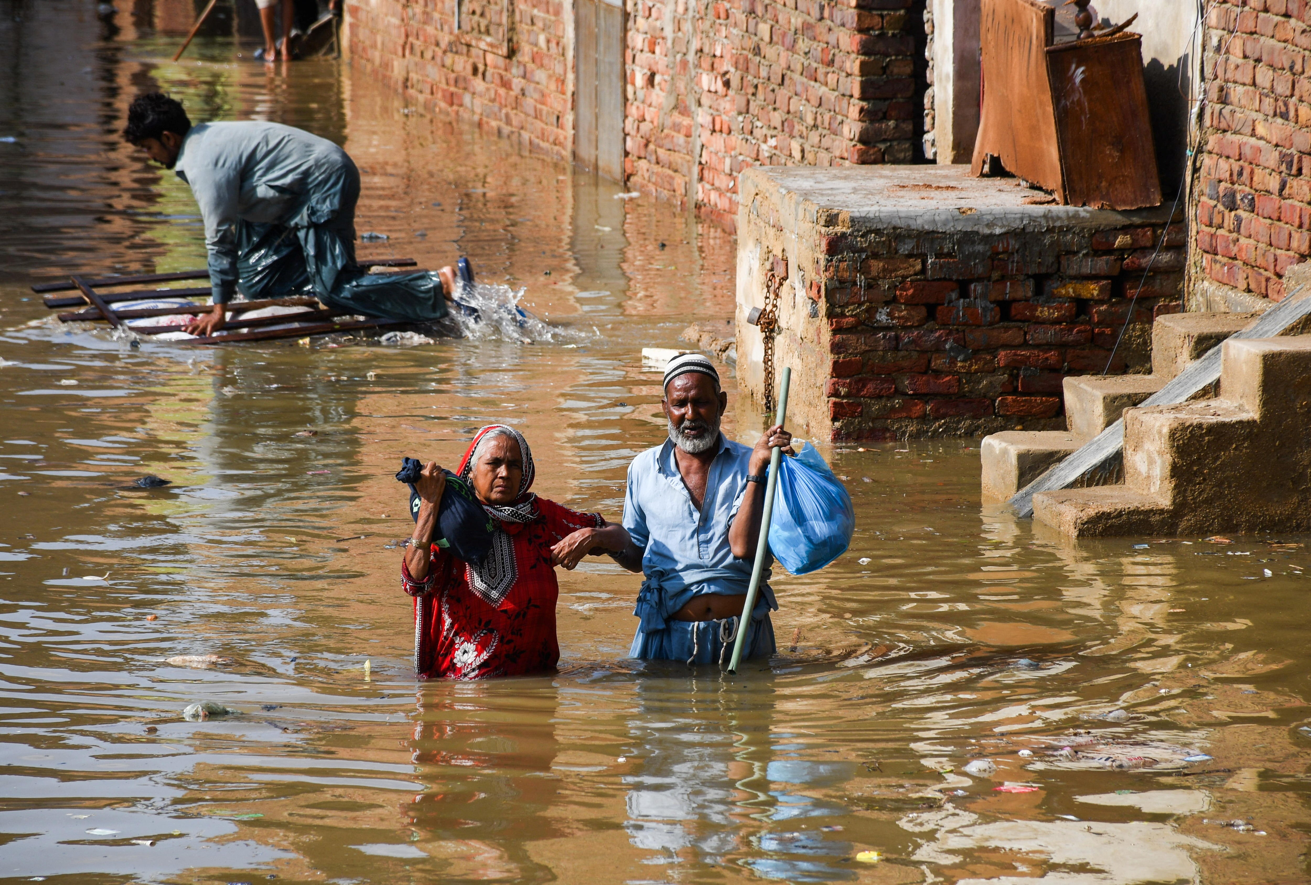 Elderly couple walk through waist-deep water next to brick houses