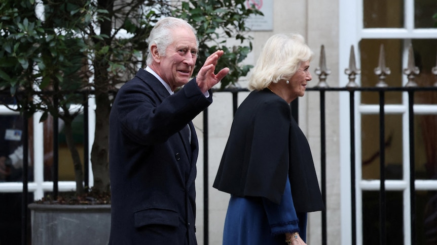 King Charles waves to a crowd as Queen Camilla walks ahead.