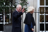 King Charles waves to a crowd as Queen Camilla walks ahead.
