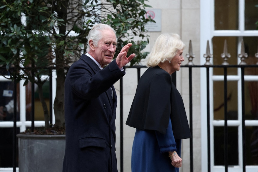 King Charles waves to a crowd as Queen Camilla walks ahead.