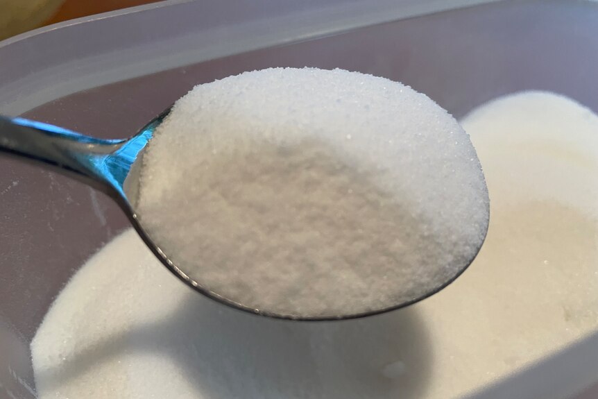 A soup spoon full of caster sugar, hovering over a container of sugar. 