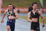 Two male runners near the line in a 100m race, with the leader's (right) hair flying in the wind.