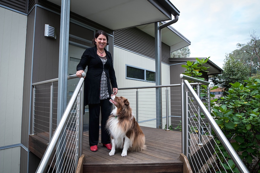 Janine Strachan stands on the elevated front porch of her dark and light grey home with brown and white border collie Duke.