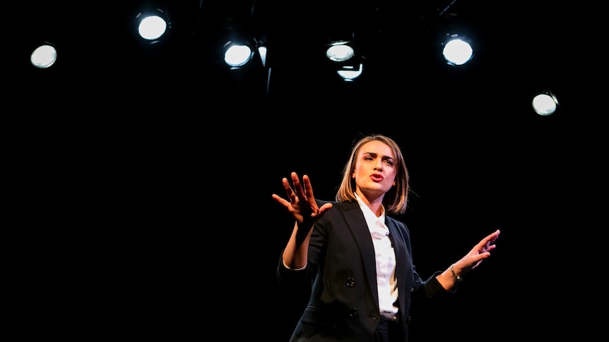 Sheridan Harbridge addresses an audience from a black stage. Stage lights are seen above her head.