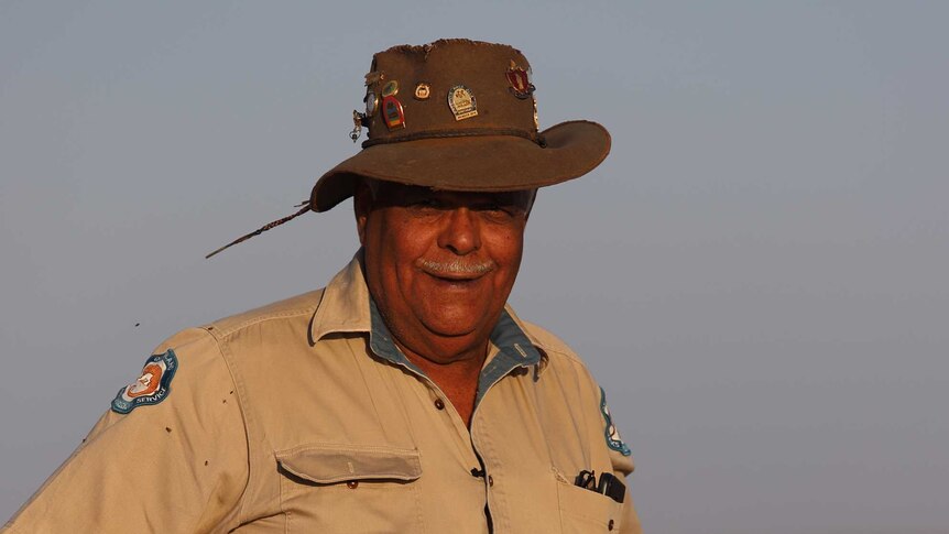 A man in an Akubra-style hat and a ranger uniform stands with hands on his hat with red dirt in the background.