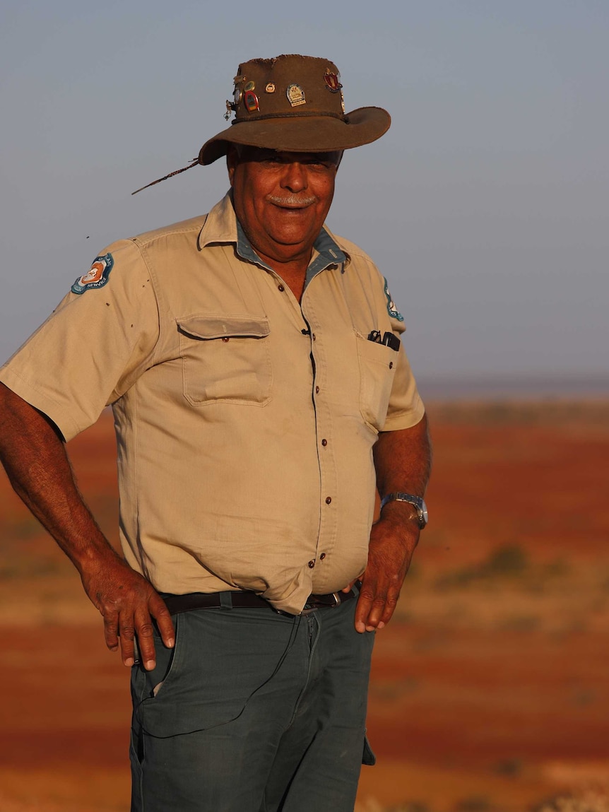 A man in an Akubra-style hat and a ranger uniform stands with hands on his hat with red dirt in the background.