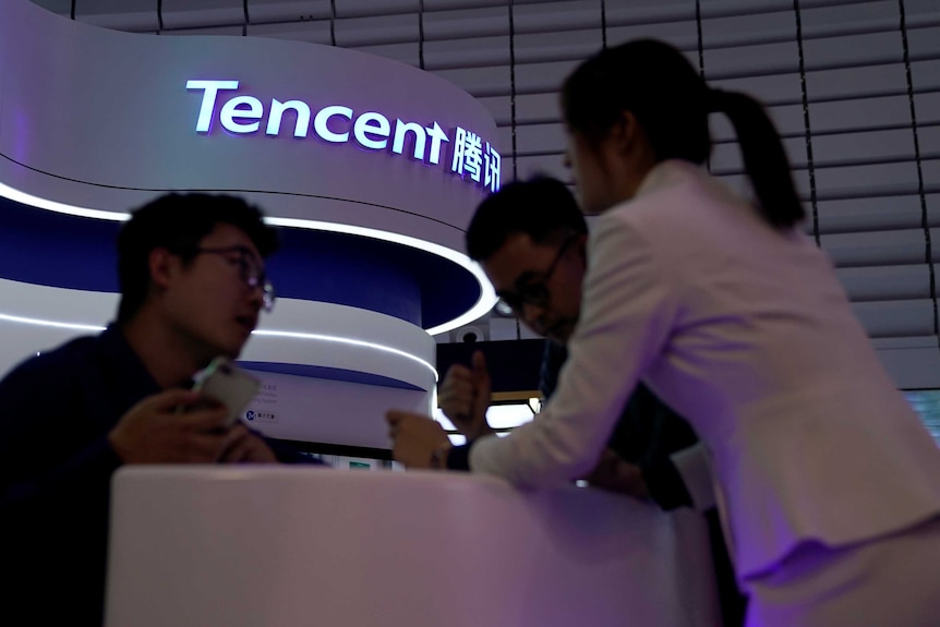 From a low angle, you look up at darkened people huddled around an information desk with a neon sign that reads 'Tencent'.
