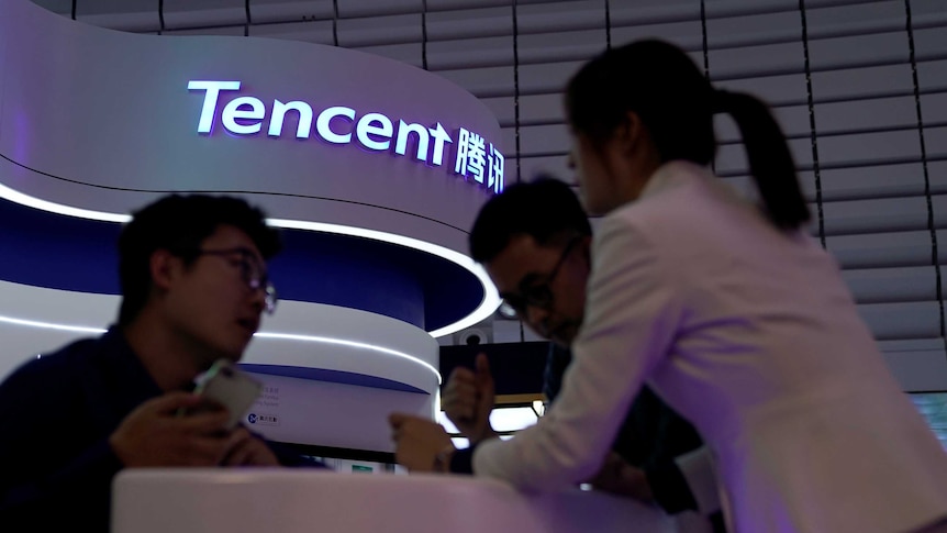 From a low angle, you look up at darkened people huddled around an information desk with a neon sign that reads 'Tencent'.