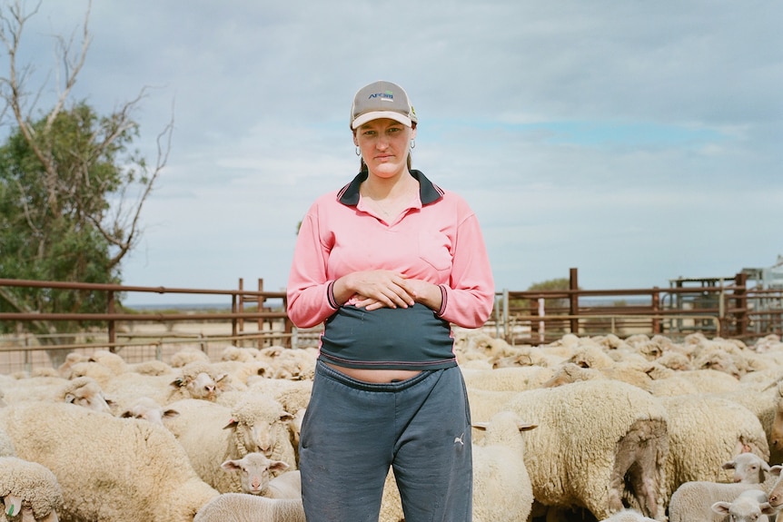 A pregnant farmer stands in a sheep pen with sheep surrounding her