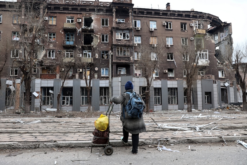 A person with a loaded trolley looks at a large apartment building that has been heavily damaged during a war.