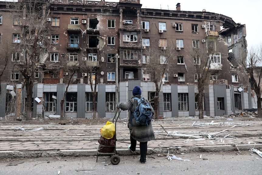 Una persona che trasporta un carro carico guarda un grande condominio che è stato gravemente danneggiato durante la guerra.