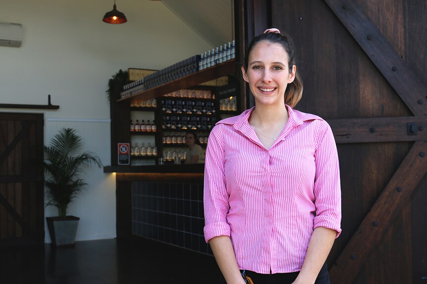 A young, slender woman in a pink shirt smiles while standing in front of a bar.