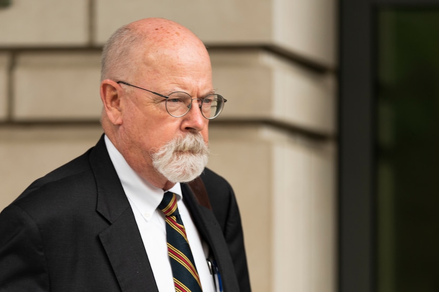 A man with bald head and white goatee and thin-rimmed round glasses appears to grimmace in black suit. 