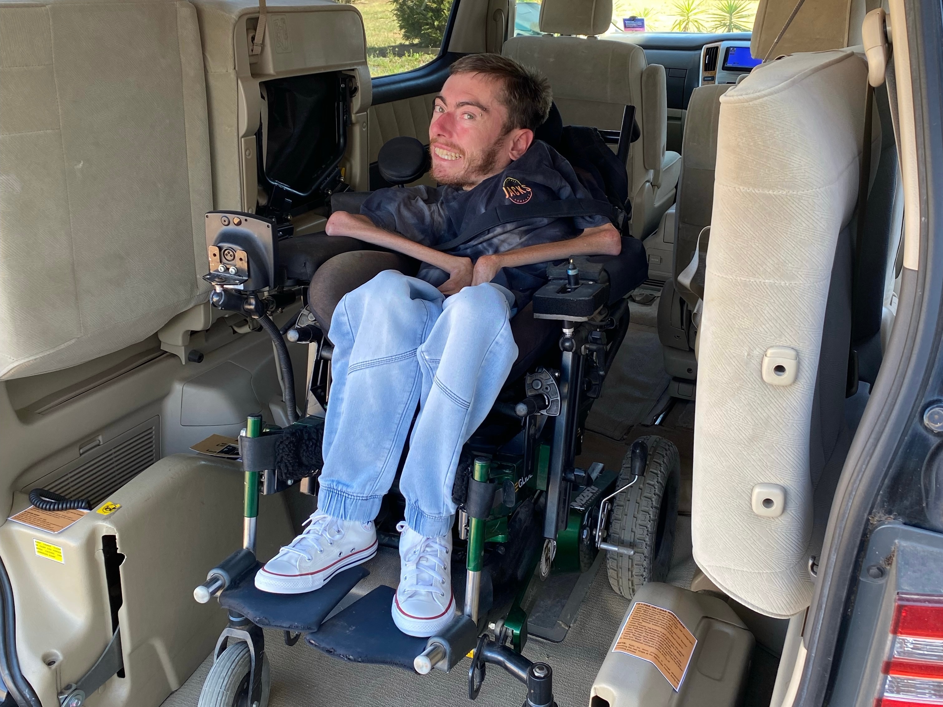 A young man in jeans sits smiling in a motorised wheelchair in the back of a disabled access van.