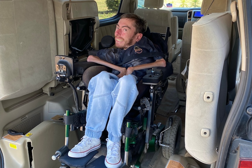 A young man in jeans sits smiling in a motorised wheelchair in the back of a disabled access van.