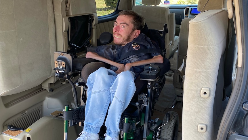 A young man in jeans sits smiling in a motorised wheelchair in the back of a accessible van.