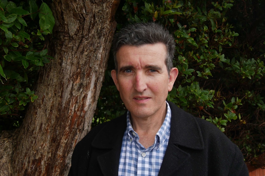 A man in a jacket and shirt  stands in front of some trees and is looking at the camera.