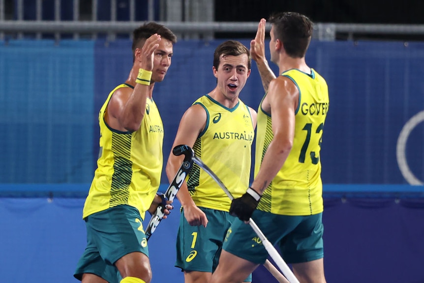 Three Australian hockey players high five each other on the field, they look excited and sweaty