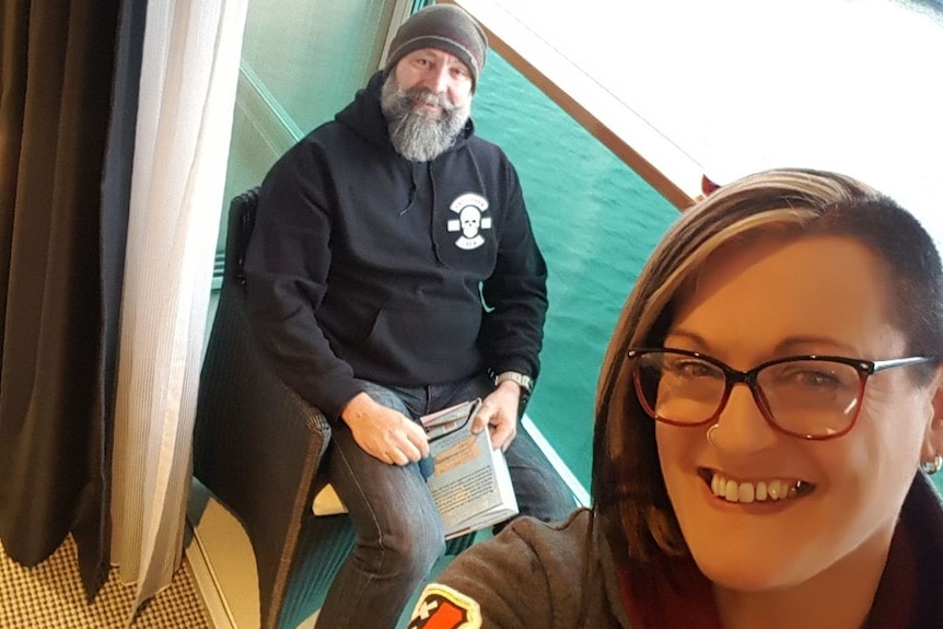 A woman takes a selfie of herself and a man on the balcony of a boat.