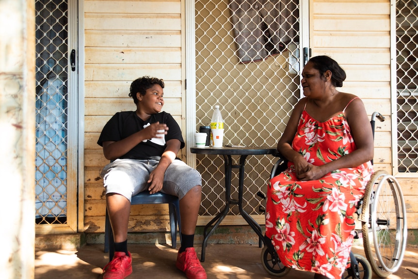15 year old Taylen sits at a round table laughing, opposite his mother, in front on a home in Katherine. 