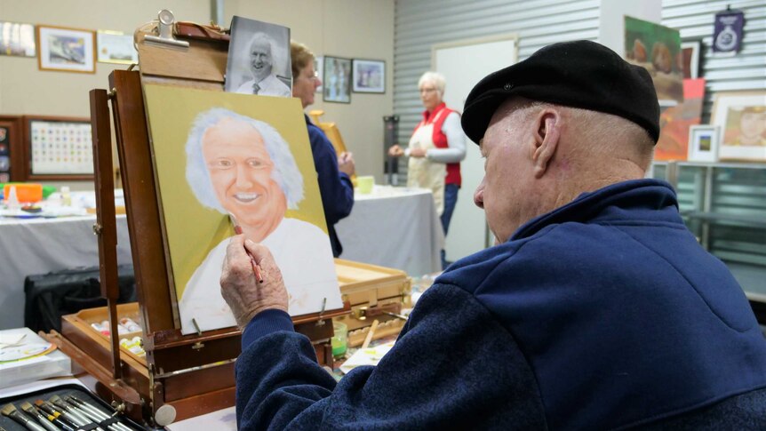 An elderly man paints at an easel.