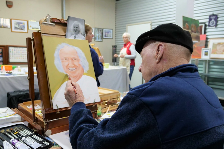 An elderly man paints at an easel.