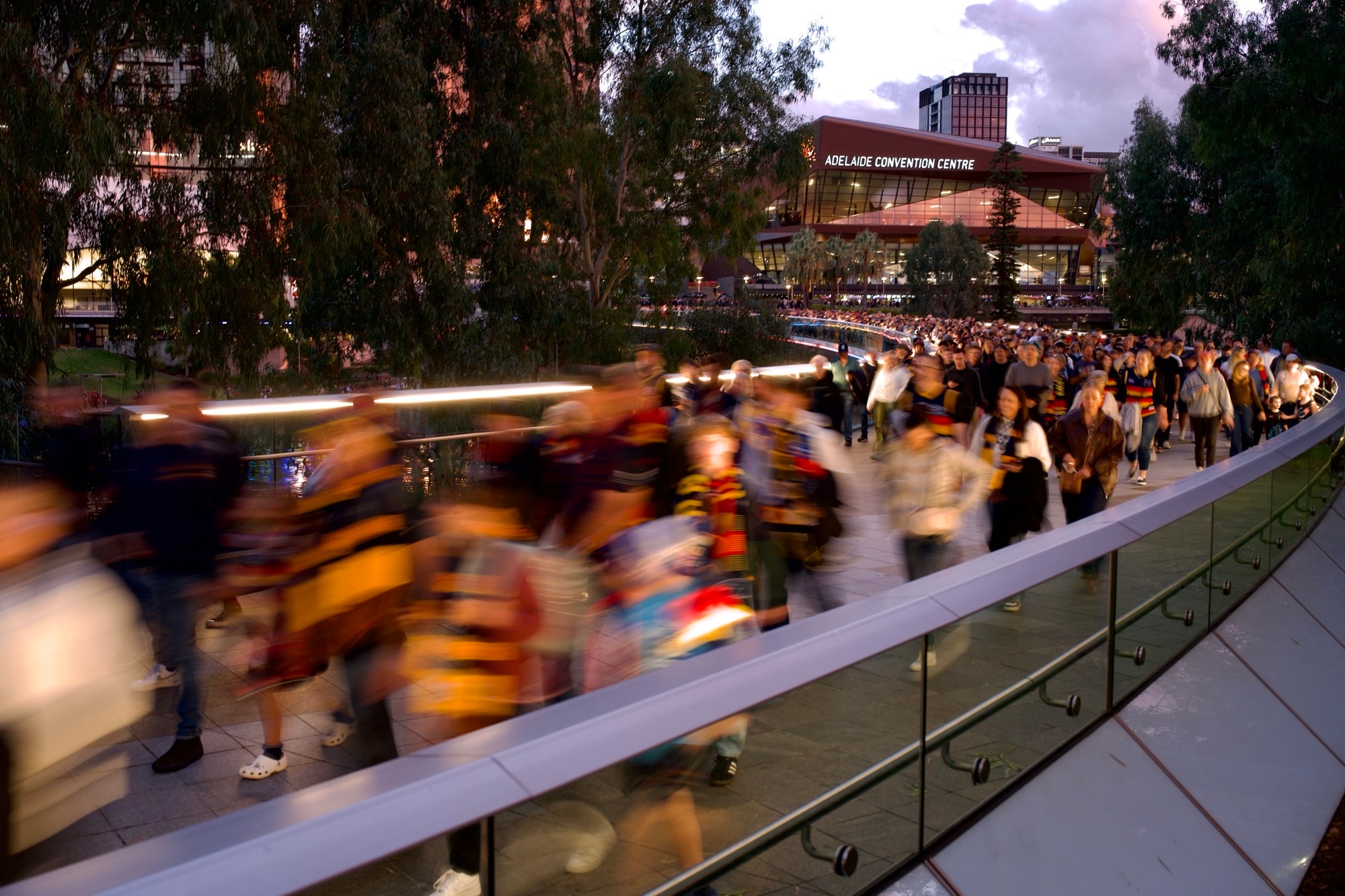 Uma imagem desfocada de longa exposição de uma multidão aglomerada atravessando uma ponte em frente ao Centro de Convenções de Adelaide. 