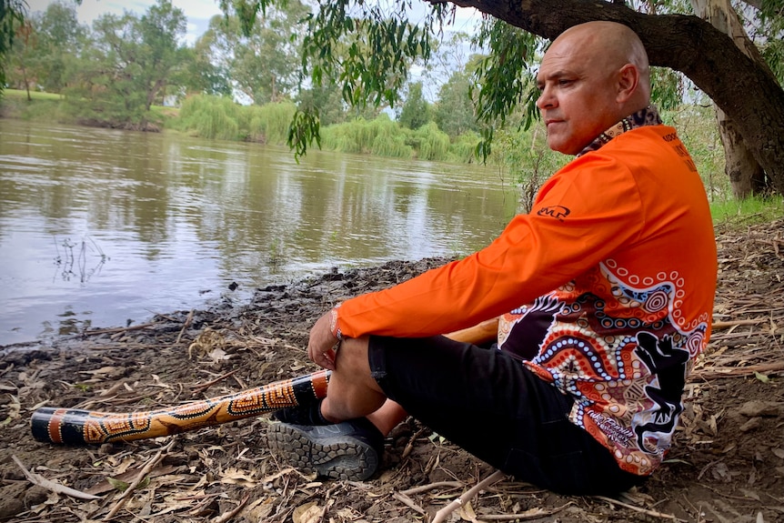Indigenous man sits on the river bank with didgeridoo