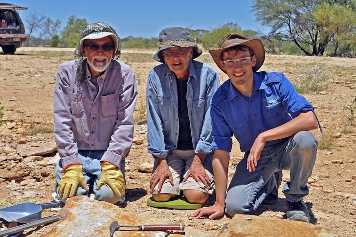 An older man and woman kneeling next to a younger fellow in the outback, fossicking tools laid out in front of them.