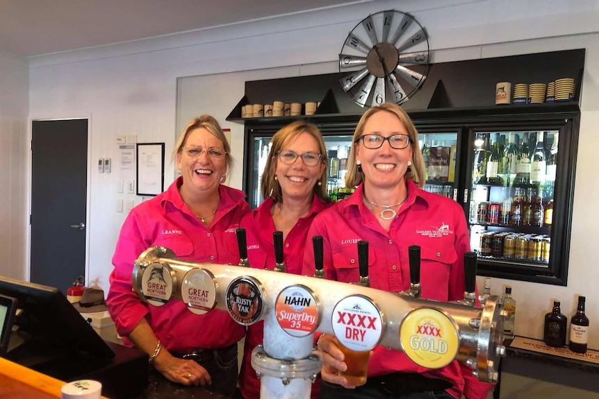 Three woman in hot pink long-sleeved shirts stand behind a beer tap at a bar.