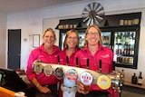 Three woman in hot pink long-sleeved shirts stand behind a beer tap at a bar.