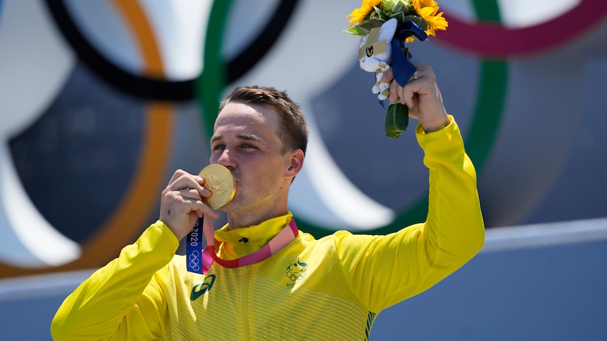 A man in a yellow jacket kisses the gold medal around his neck. He also holds a small bouquet of flowers in the air.