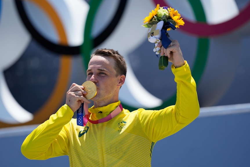 A man in a yellow jacket kisses the gold medal around his neck. He also holds a small bouquet of flowers in the air.