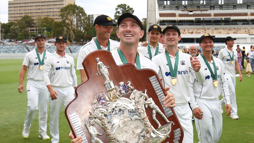 A happy man carries a big cricket trophy surrounded by teammates