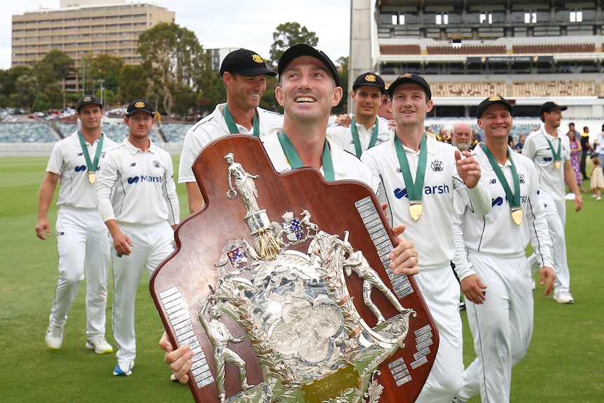 A happy man carries a big cricket trophy surrounded by teammates