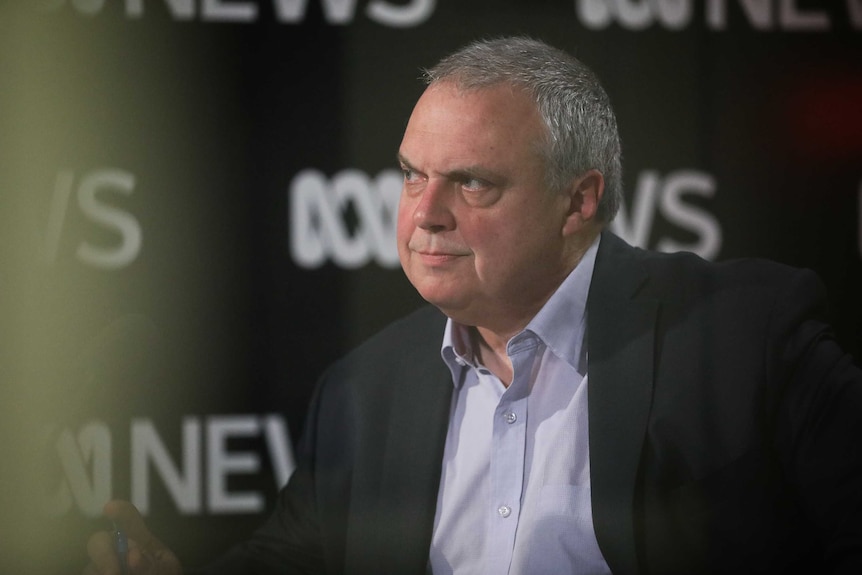 Griff is in a radio studio, wearing a suit and no tie, looking angrily right to left.