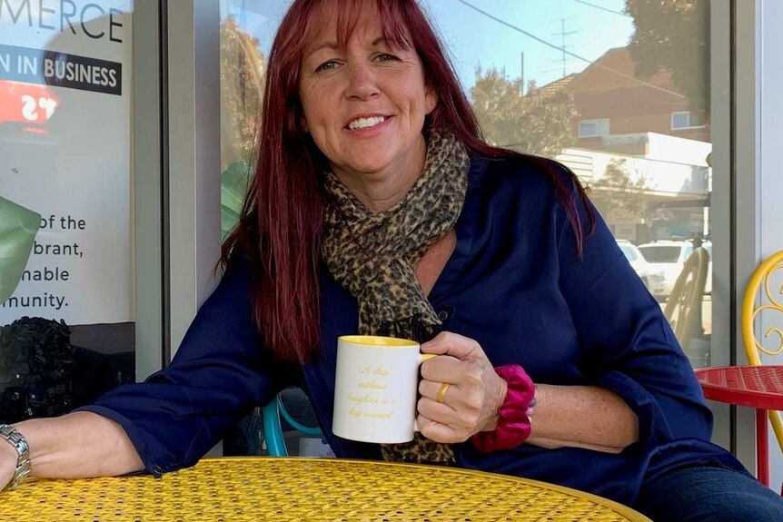 Photo of Esther Bliss, holdling a mug sat at a yellow cafe style bench in front of a window.