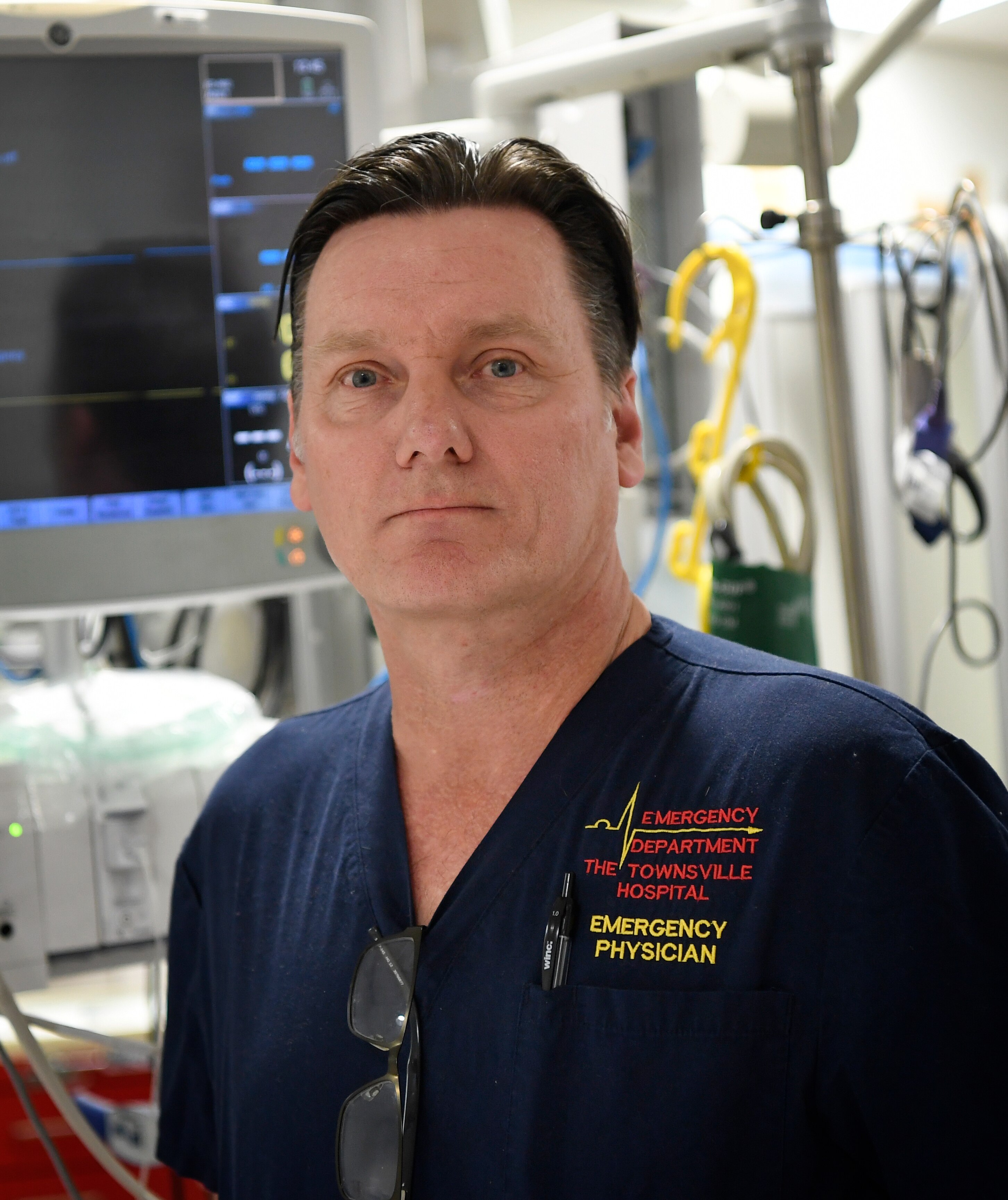 A serious looking middle-aged man with slicked back hair, standing in hospital ward with equipment, wearing blue scrubs.