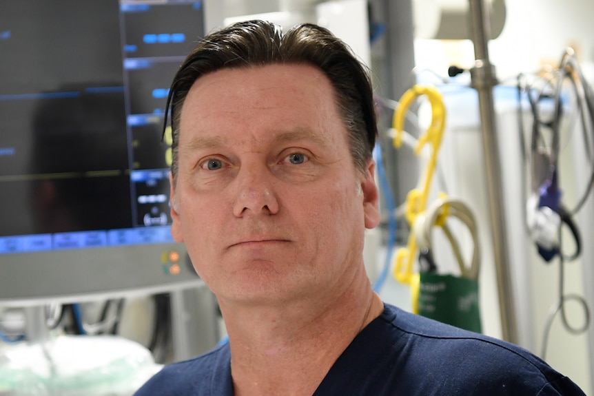 A serious looking middle-aged man with slicked back hair,  standing in hospital ward with equipment, wearing blue scrubs.