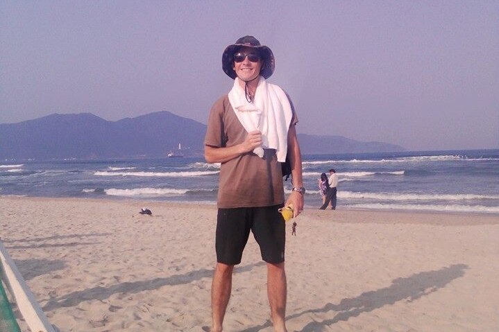 A man wearing a hat stands on a beach with a towel on his shoulders
