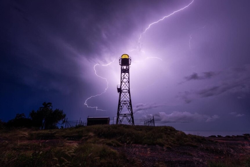 Lightning splits the night sky, illuminating a small lighthouse in the foreground.