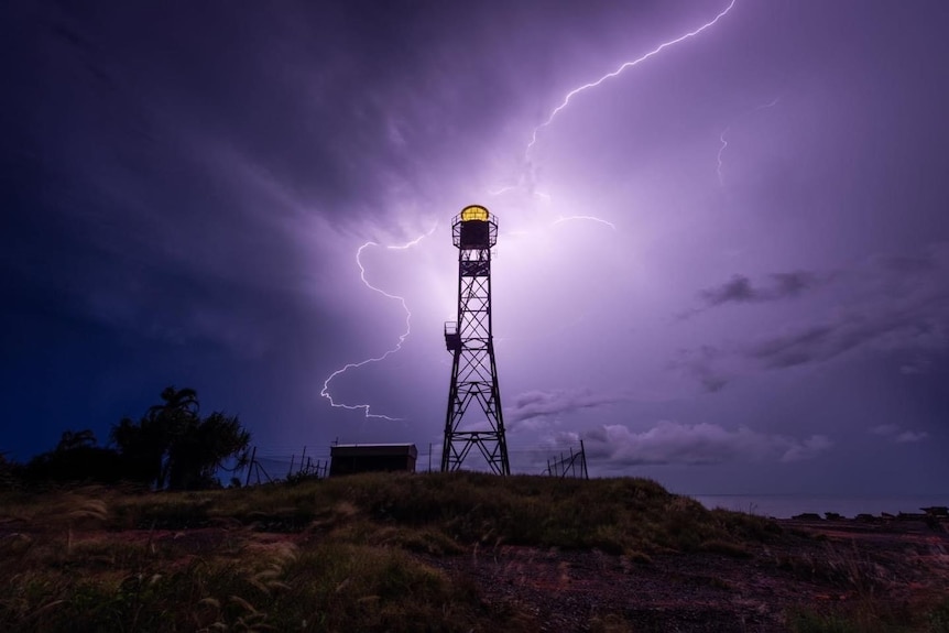 Lightning splits the night sky, illuminating a small lighthouse in the foreground.