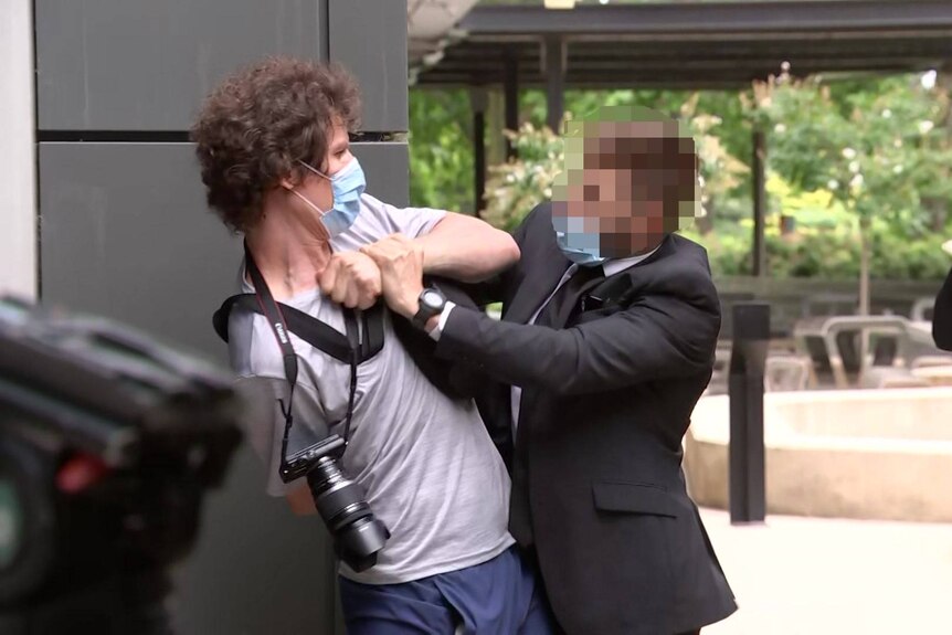 A man wearing a mask with a camera around his neck has his arms restrained by a man in a black suit