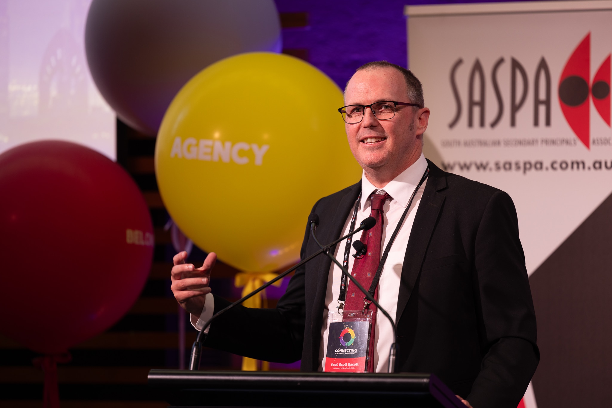 Homem atrás do pódio, fazendo um discurso, com um balão da agência atrás dele.