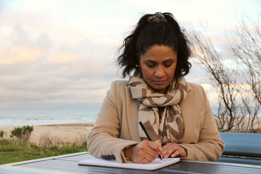  Woman in coat and scarf writes on a piece of paper on a bench by the beach