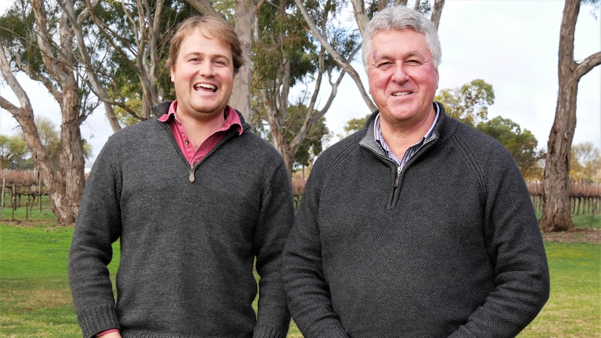 A younger and an older man grin. They're standing in front of gum trees and behind the trees is a vineyard 