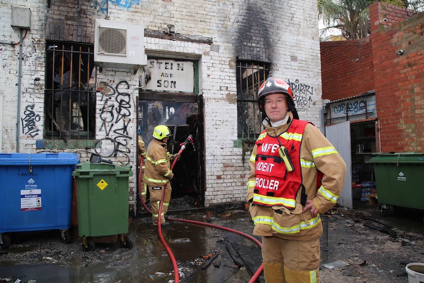 A firefighter stands in the foreground while in the background another firefighter sprays water on the burnt building.