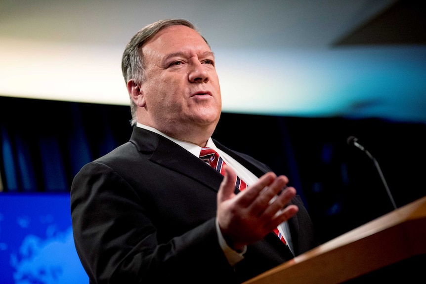 A man wearing a dark suit with a red tie stands at a podium gesturing with his hand.