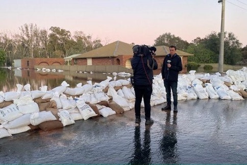 Cameraman filming reporter holding microphone standing next to row of sandbags in residential street flooded on other side.